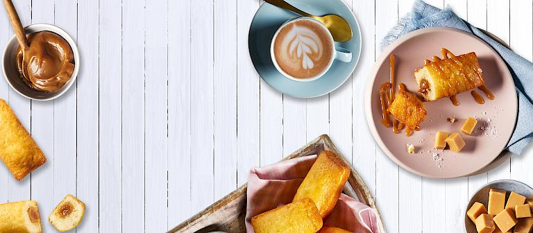 Einige Mini Cakes mit Karamellfüllung arrangiert mit flüssigem und festem Karamell auf einem Teller daneben eine Tasse Cappucino.