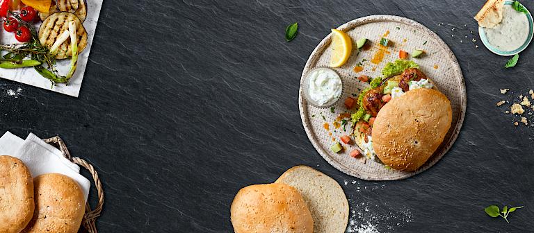 A mini flatbread sandwich on a light-colored plate, a sliced flatbread, a plate of grilled vegetables, yogurt dip, herbs, and a lemon wedge, accompanied by additional flatbreads in a basket and small bowls of dip. All set against a dark slate background.
