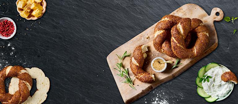 A top view of several golden-brown simit rings sprinkled with sesame seeds on a wooden board against a dark stone background, accompanied by small bowls of dip, spices, cucumber slices, and fresh herbs.