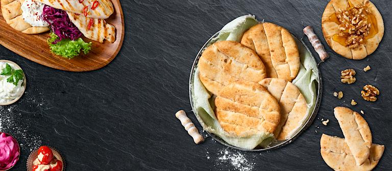 A top view of several light brown mini pita breads baked on a dark stone surface, accompanied by serving suggestions featuring stuffed pita, dips, nuts, and both sweet and savory toppings.
