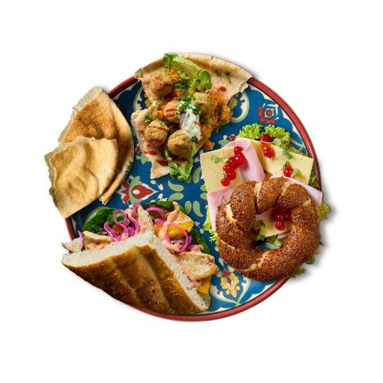 A round serving platter featuring an assortment of Middle Eastern dishes: on the left, flatbread and a sandwich with meat, salad, and pickled vegetables; at the top, falafel in flatbread; on the right, a simit ring with cheese and cold cuts next to it, all arranged on a decoratively patterned plate.