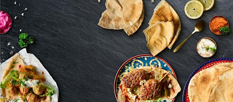 Arabic flatbreads with grilled meat skewers, dips, and side dishes arranged on decorative plates against a dark slate background, surrounded by lemon slices and fresh herbs.