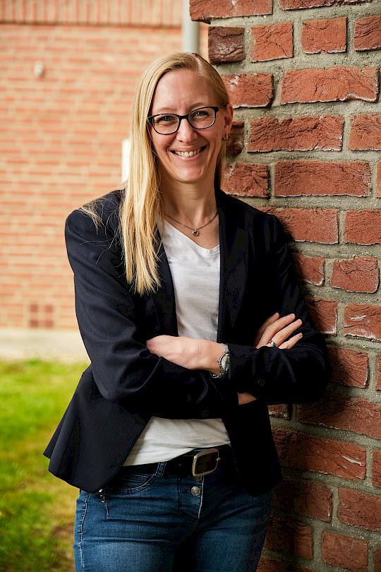A woman with long blonde hair wears a black blazer and white shirt. The background includes plants.