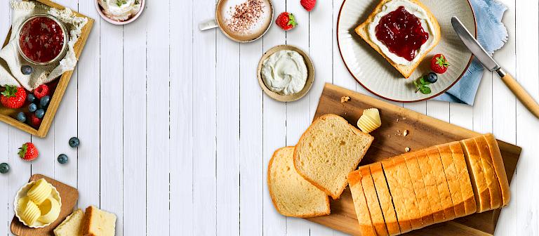 Breakfast scene on a white wooden table: sliced brioche on a wooden board with butter, next to it a slice with jam and cream, fresh berries, a jar of jam, and a cup of cappuccino.