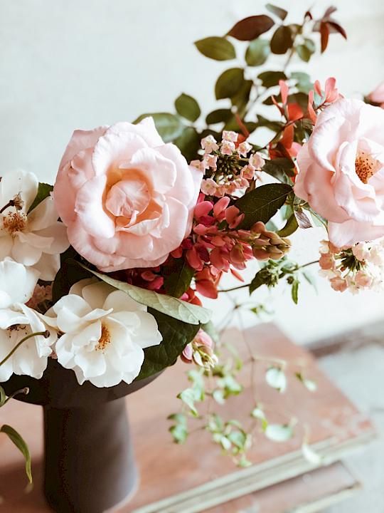 Flower arrangement with pale pink roses, white blossoms, pink buds, and green branches in a dark vase against a neutral, blurred background.