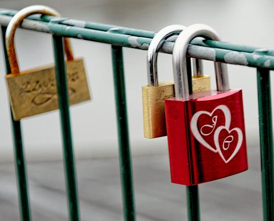 Three padlocks on a green metal railing; two gold-colored, one red with engraved hearts and the letters.