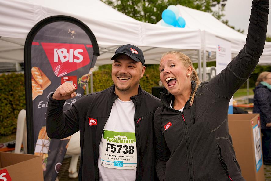 Two people at a company run wearing IBIS jackets; one wears a T-shirt with race number and company name. Background includes white tents, blue balloons, and IBIS advertising banner.
