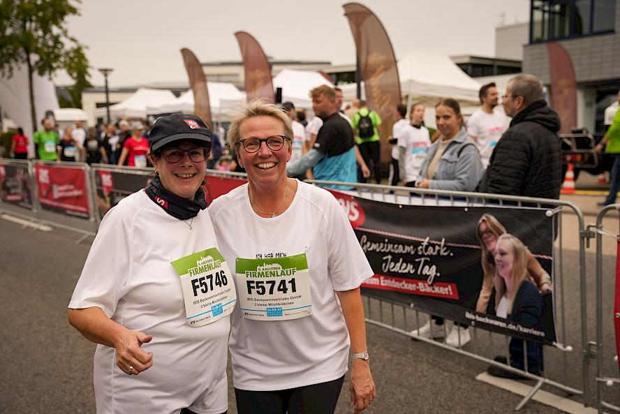 Two people wearing white shirts with corporate run bib numbers F5746 and F5741 at outdoor event; other participants, tents, banners, and fencing in the background.