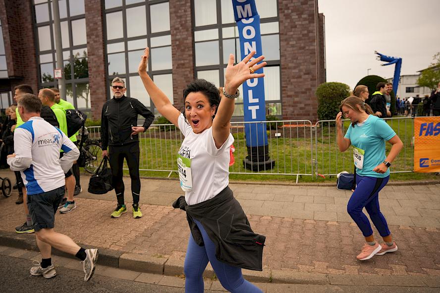 Person wearing white shirt raises arms at running event; other participants, spectators, bicycles, “MONOLIT” banner, and building with large windows in the background.