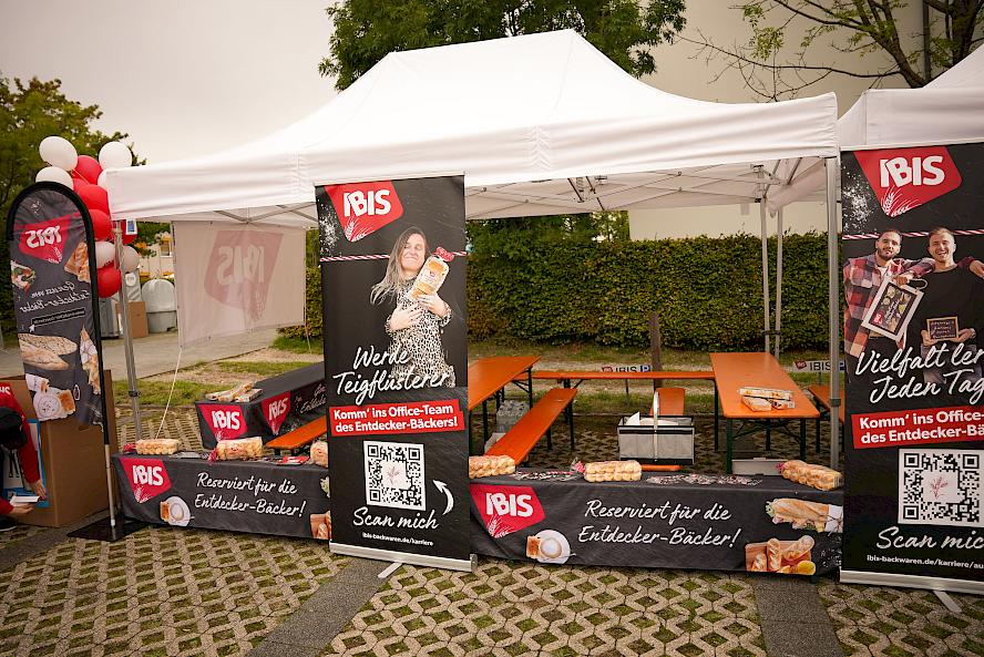 Exhibition booth with white pavilion, IBIS banners showing bread products and QR codes; tables with baked goods, red and white balloons on the left side.