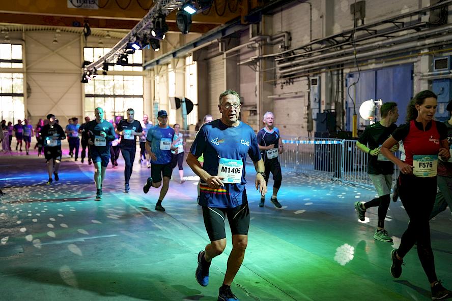 Group of people in athletic clothing running in a hall with high ceilings, large windows, and lighting; metal barriers along the running path.
