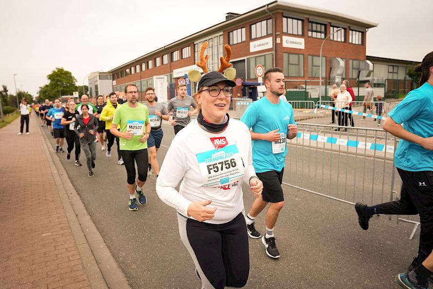 Street run with several participants in athletic clothing; person in white shirt with reindeer antler headband in foreground; barriers and spectators in the background.