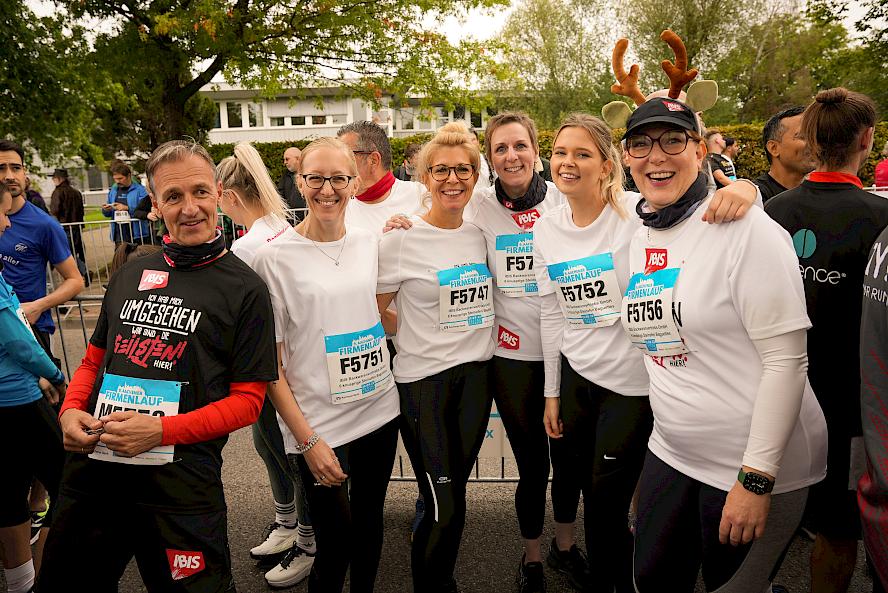Group of people in athletic clothing at a running event; white T-shirts with “Firmenlauf” and “IBIS” print; other participants, trees, and a building in the background.